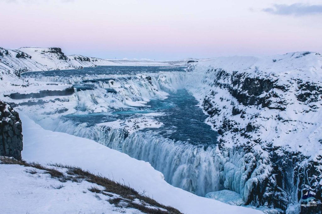 View of the Gullfoss waterfall frozen in winter, ground covered in thick snow, and pink skies.