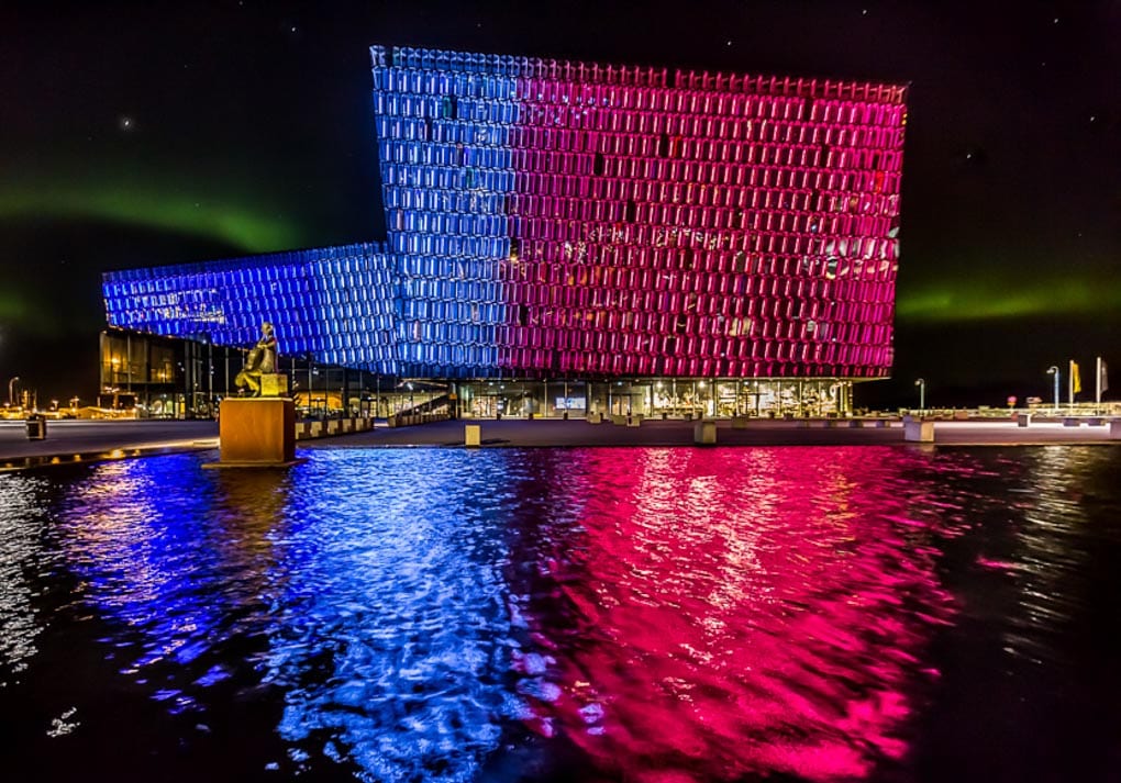 Harpa Concert Hall in Reykjavík lit in blue and pink at night, with colorful reflections on the water.
