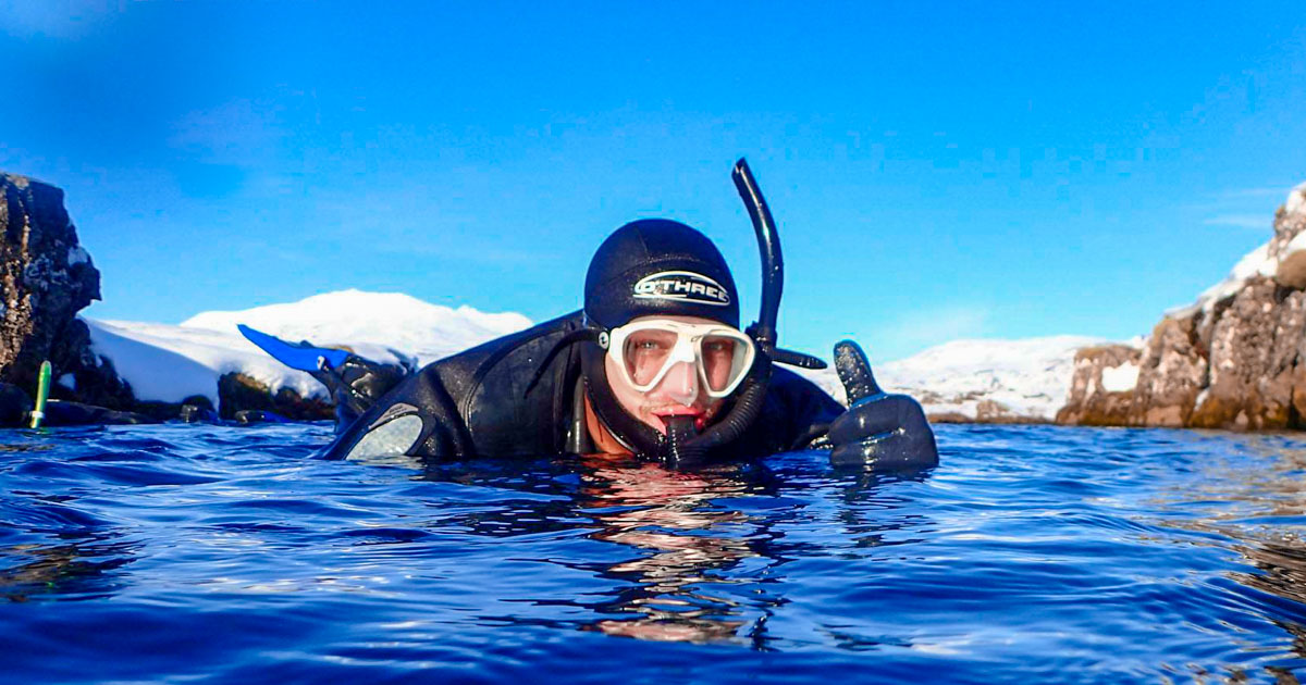 Close up of man in snorkelling suit and goggles in deep blue sea at Silfra Fissure in Iceland, showing a thumbs up.