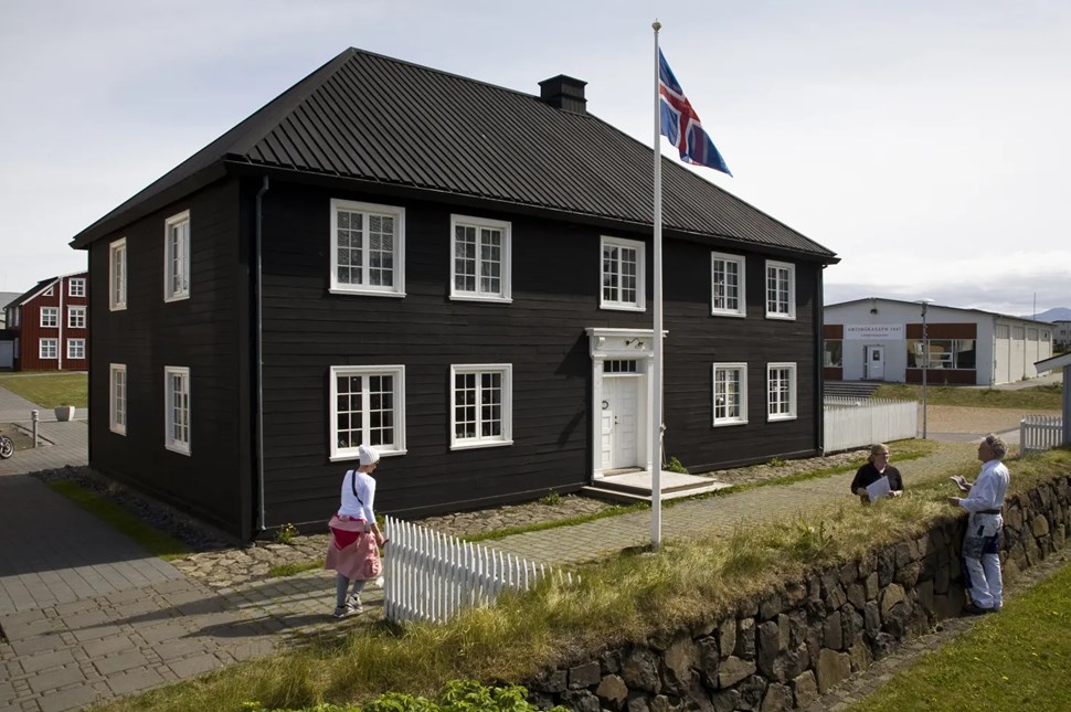 Icelandic black wooden house with an Icelandic flag and three individuals outside by stone wall