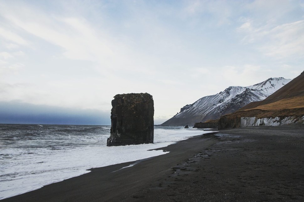 Rugged coastal landscape with towering rock formation, snowy mountains, and dark sandy shores