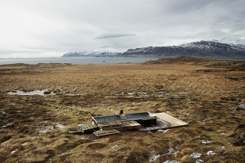 Person soaking in outdoor tub amidst vast Icelandic tundra with snow-capped mountains in the distance