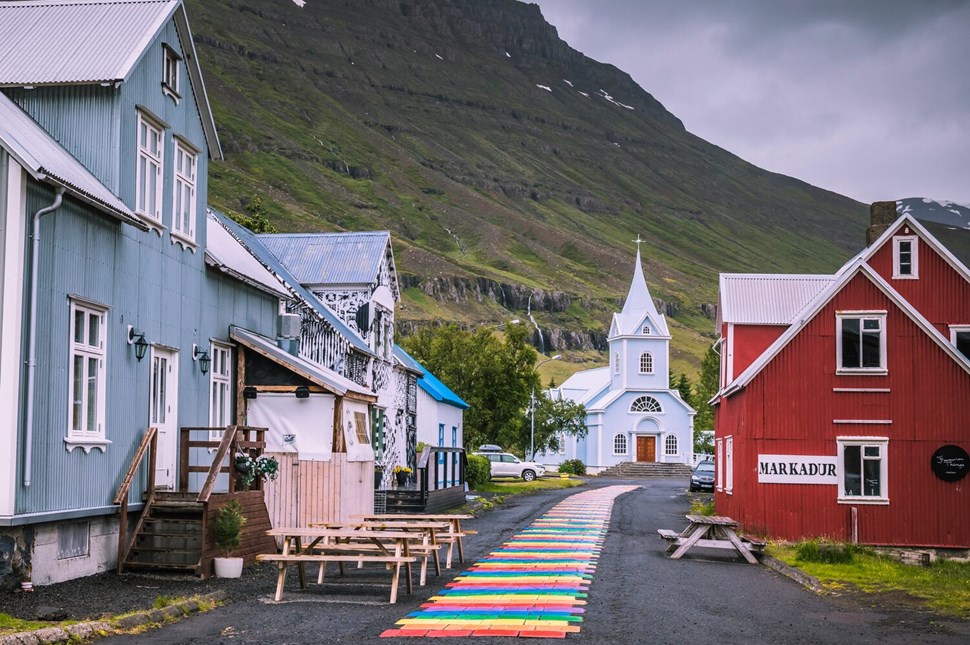 Quaint village street with colorful houses, rainbow pathway, and white church against mountain backdrop