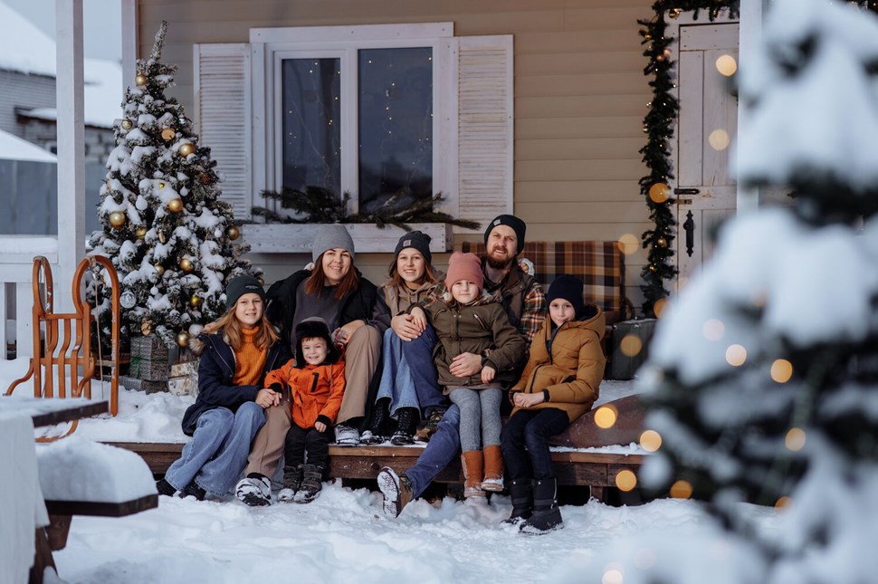 Family sits together on snowy porch, flanked by decorated Christmas trees, embracing holiday spirit.
