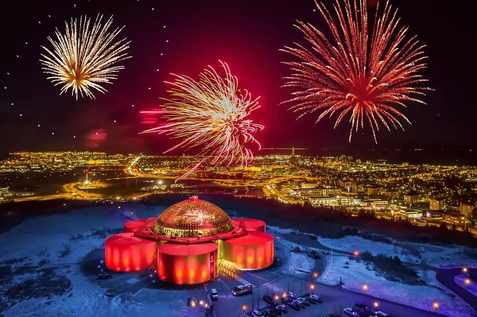 Brilliant fireworks display over illuminated Perlan Museum with Reykjavik cityscape in background.