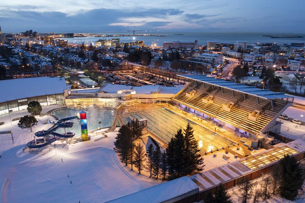 Twilight view of illuminated geothermal pool complex in snowy Reykjavik with city and ocean in background.