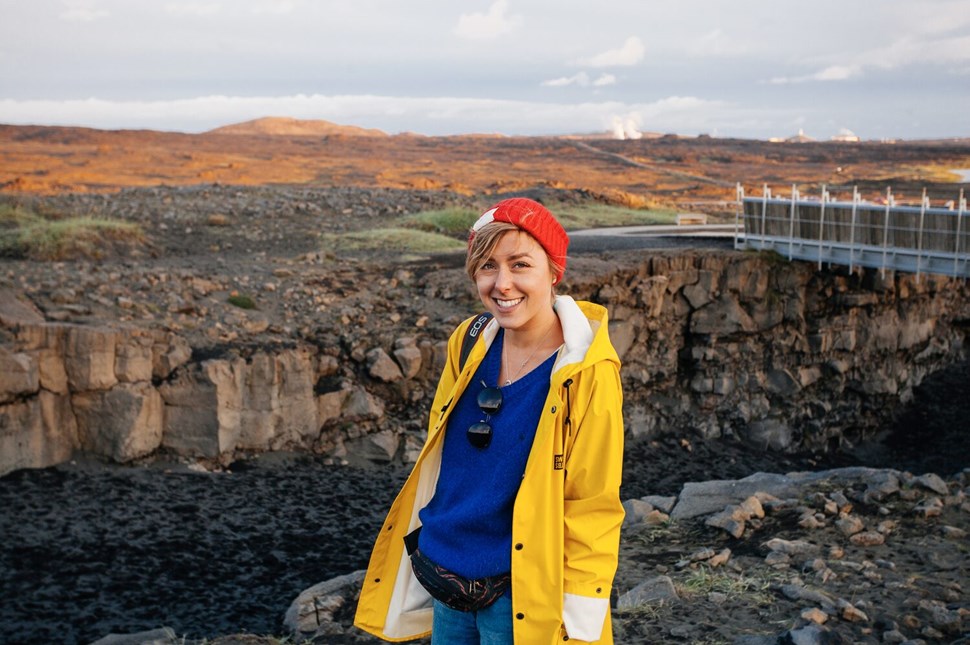Smiling woman in yellow raincoat standing by volcanic rift in Iceland, with bridge in the background.