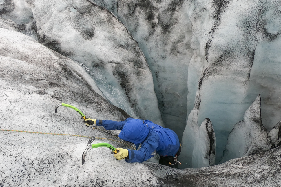 A person in a blue coat with outstretched arms holding ice axes climbing up from a hole in a glacier in Iceland