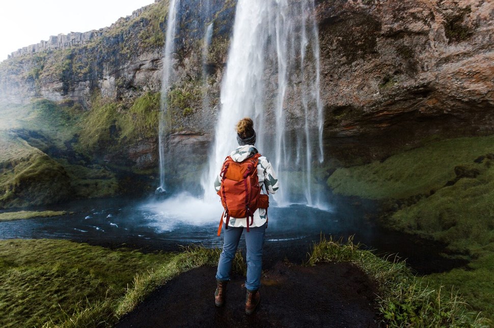 Woman with orange backpack stands facing waterfall in Iceland, mist rising around her as water cascades down lush, green cliffside.