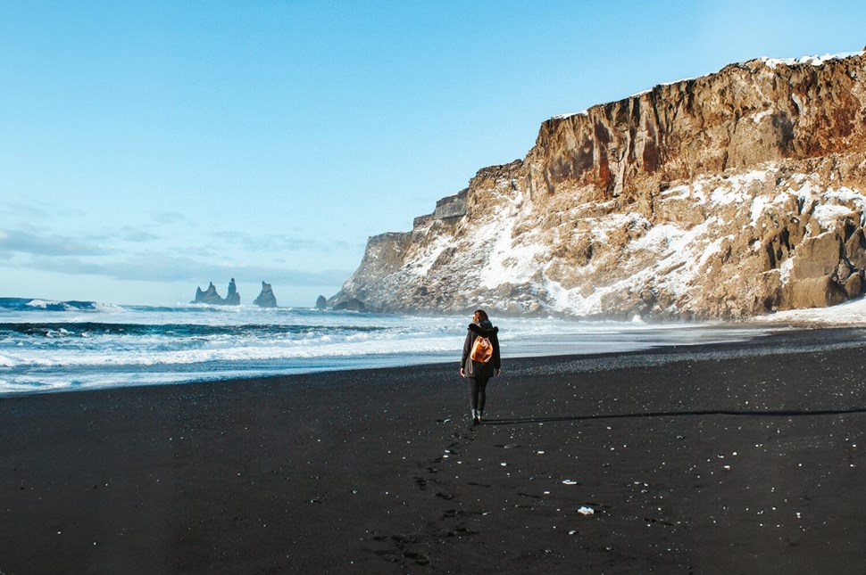 Lone traveler walks along famous Reynisfjara Black Sand Beach in Iceland, with its stark cliffs and distinctive sea stacks visible in distance.