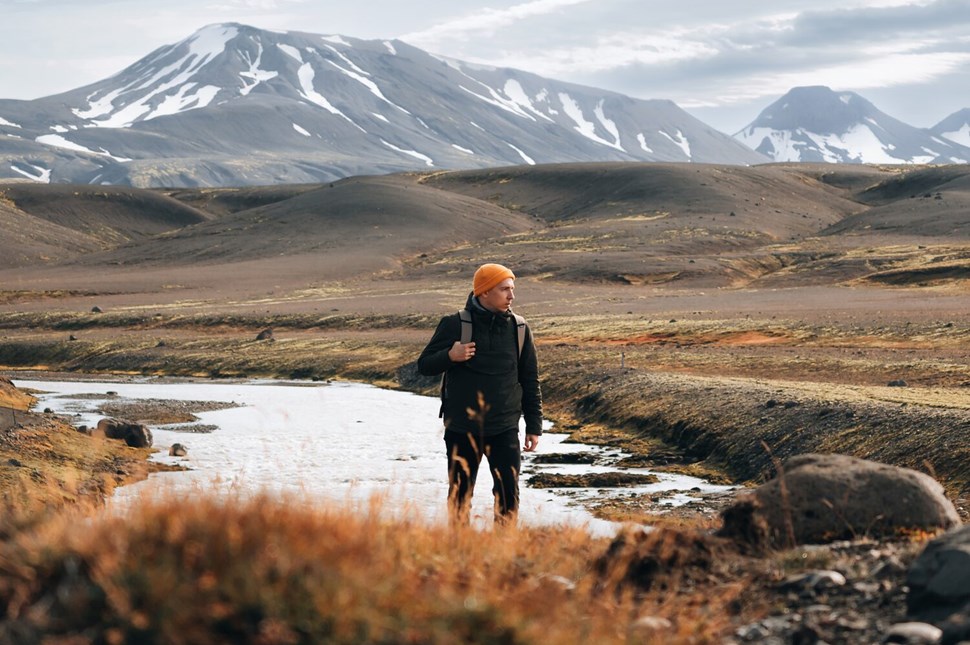 Person with an orange beanie hikes through an open field in Iceland, with snow-capped mountains in the background.