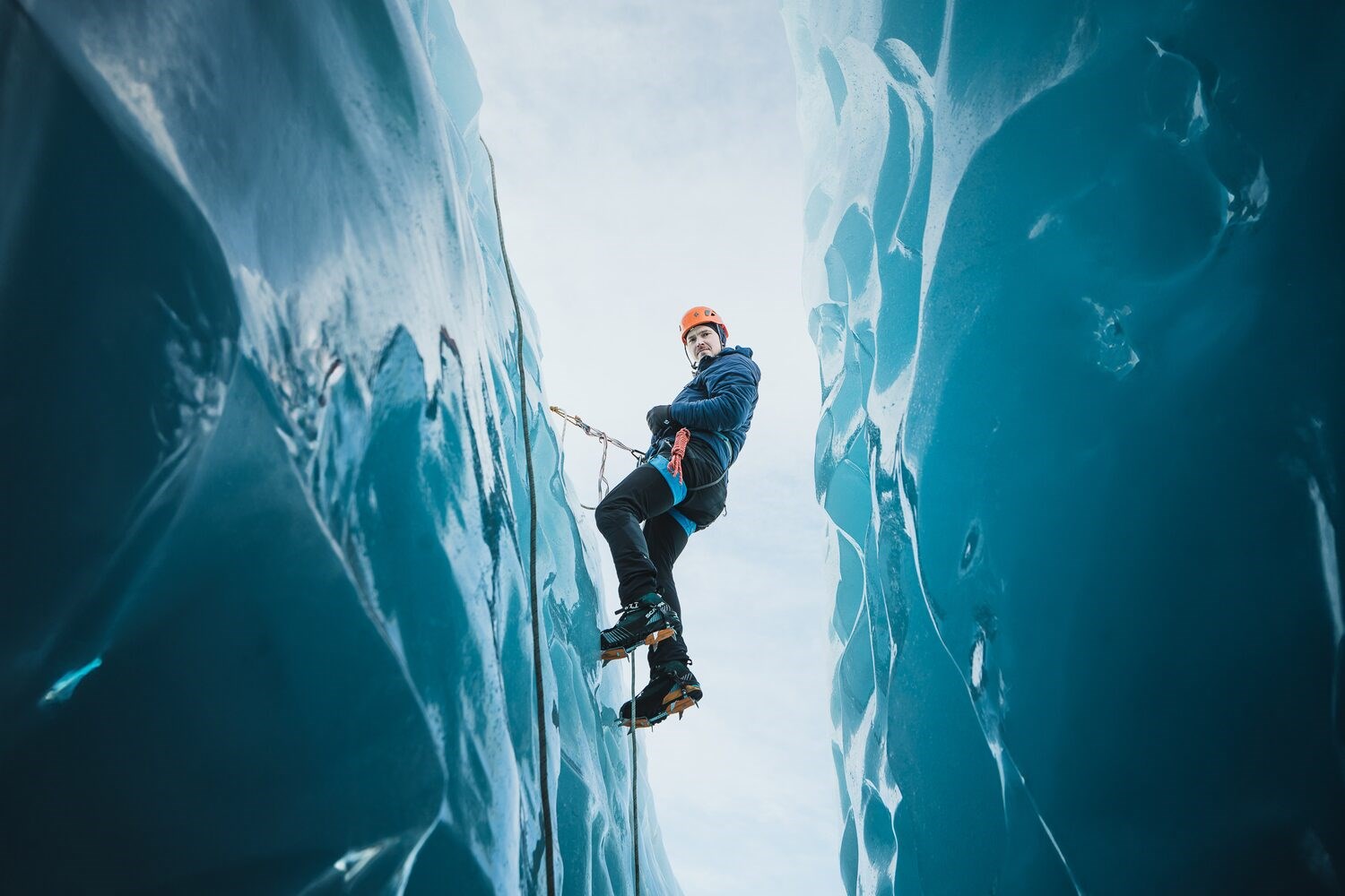 Climber ascends a glacial crevasse in Skaftafell, Iceland, equipped with ice axes and crampons against a backdrop of brilliant blue ice.
