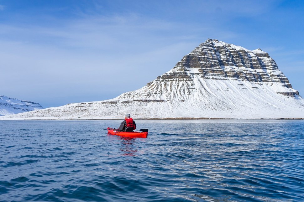 Kayaker in a red kayak and life jacket paddles in the waters near the snow-covered Kirkjufell mountain in Iceland.