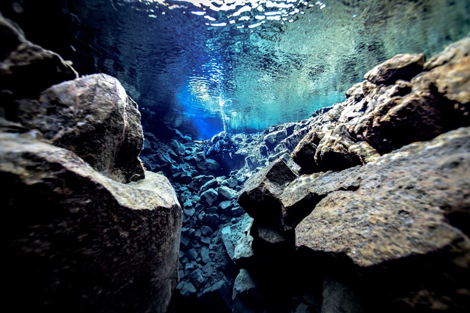 Diver explores the crystal-clear waters of the Silfra fissure in Iceland, floating between rocky formations with light filtering through the surface above.