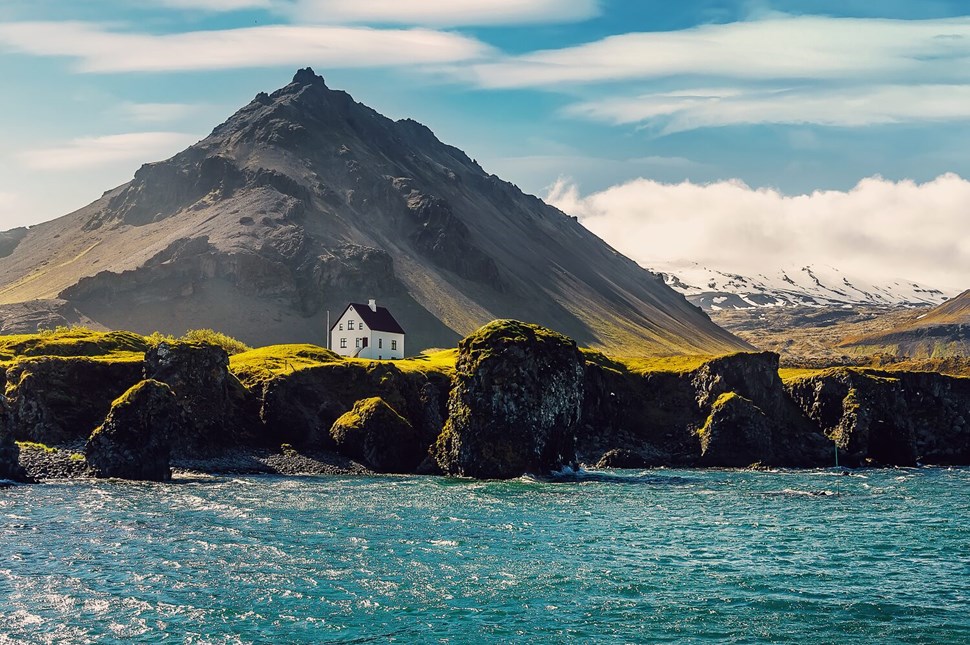 Solitary white house with a dark roof stands out against the mossy green cliffs and rough waters of Iceland's Snaefellsnes Peninsula, with a majestic mountain backdrop.