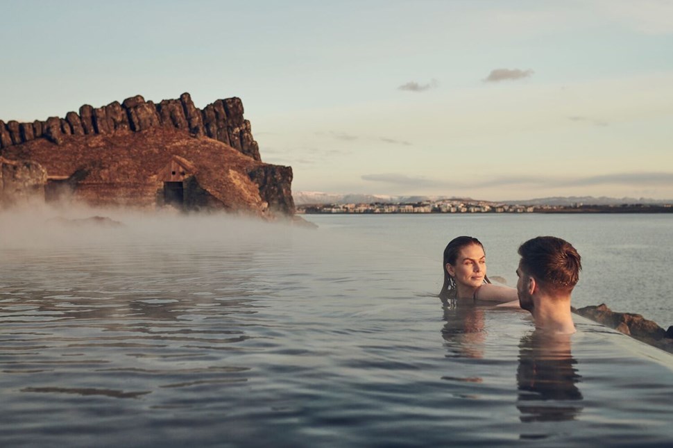 Couple enjoys a serene dip in the geothermal waters of Sky Lagoon, with mist rising around them and a rugged cliff backdrop, under a soft sunset sky.