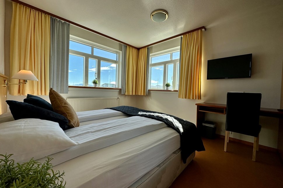 Cozy hotel room with a double bed, flanked by gold and grey curtains, showcasing a bright window view and a wall-mounted TV.