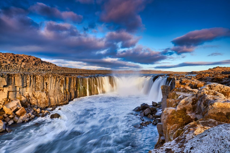 Rushing waters of waterfall cascade down cliffs under a sky streaked with pink and blue hues at sunset in Iceland.