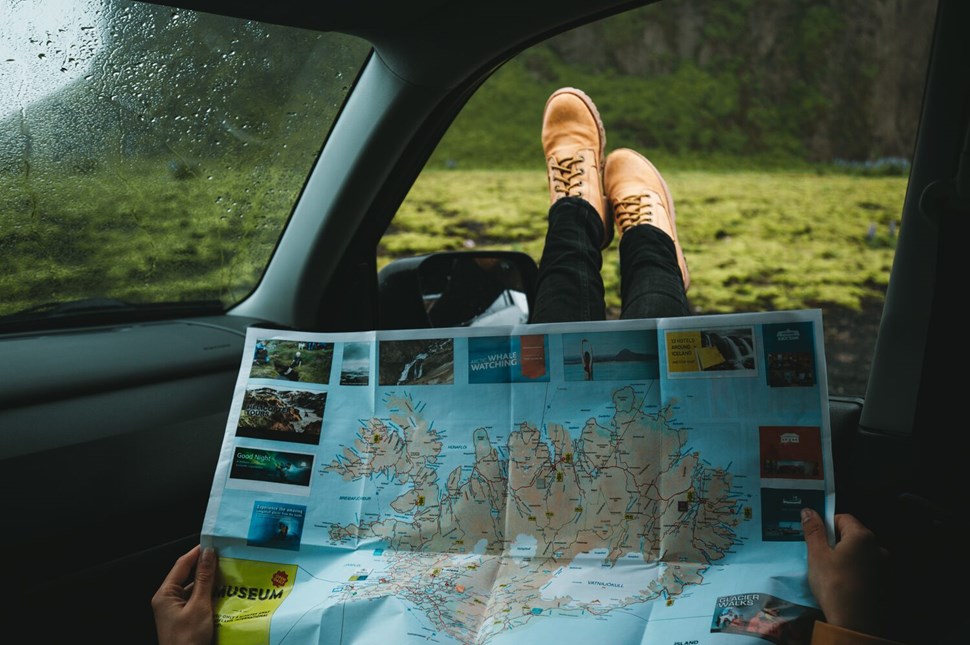 Person's feet up on a car dashboard holding a map of Iceland, with the lush Icelandic landscape visible through the rain-speckled window.