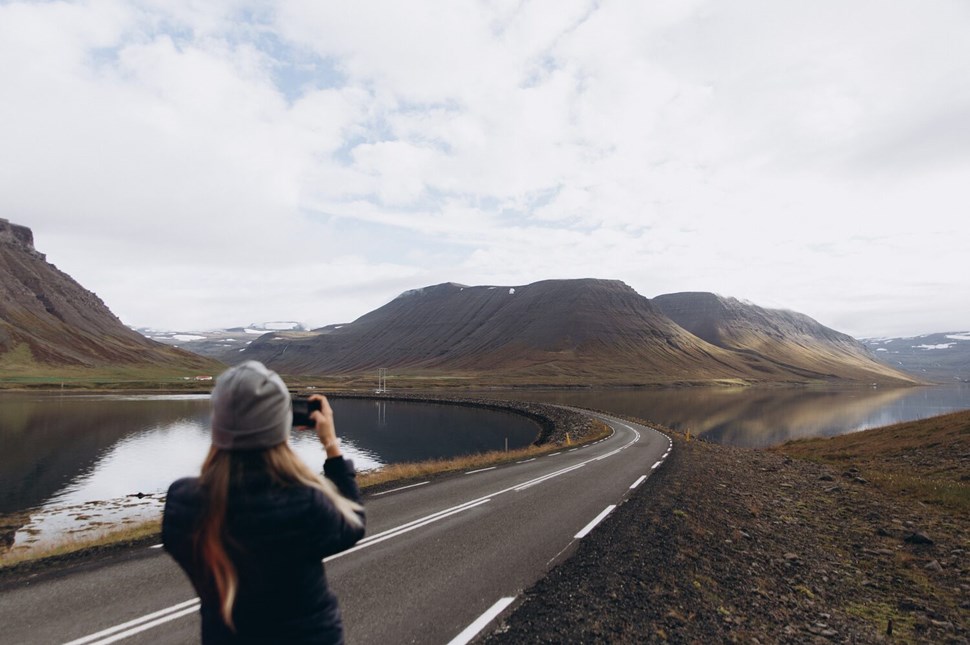 Traveler taking a photo of a picturesque road winding between calm waters and mountainous landscapes in Iceland.