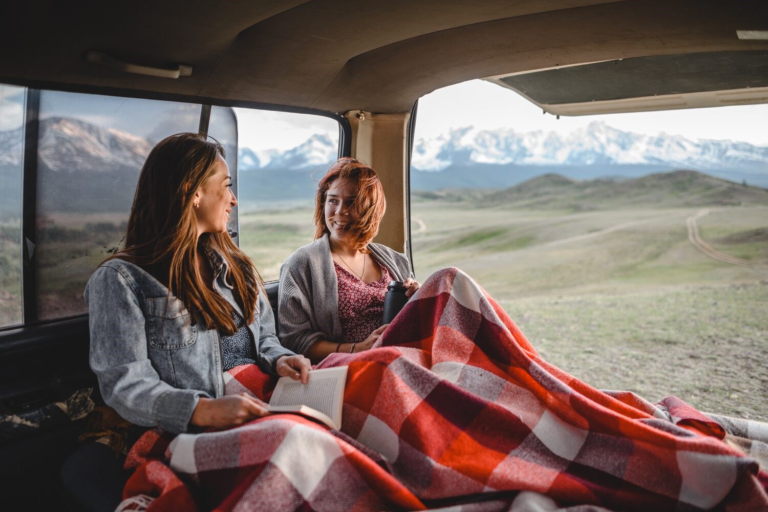 Two women in back of camper van, in red tartan blanket, smiling with books and flask, view of snowy Iceland mountains in background