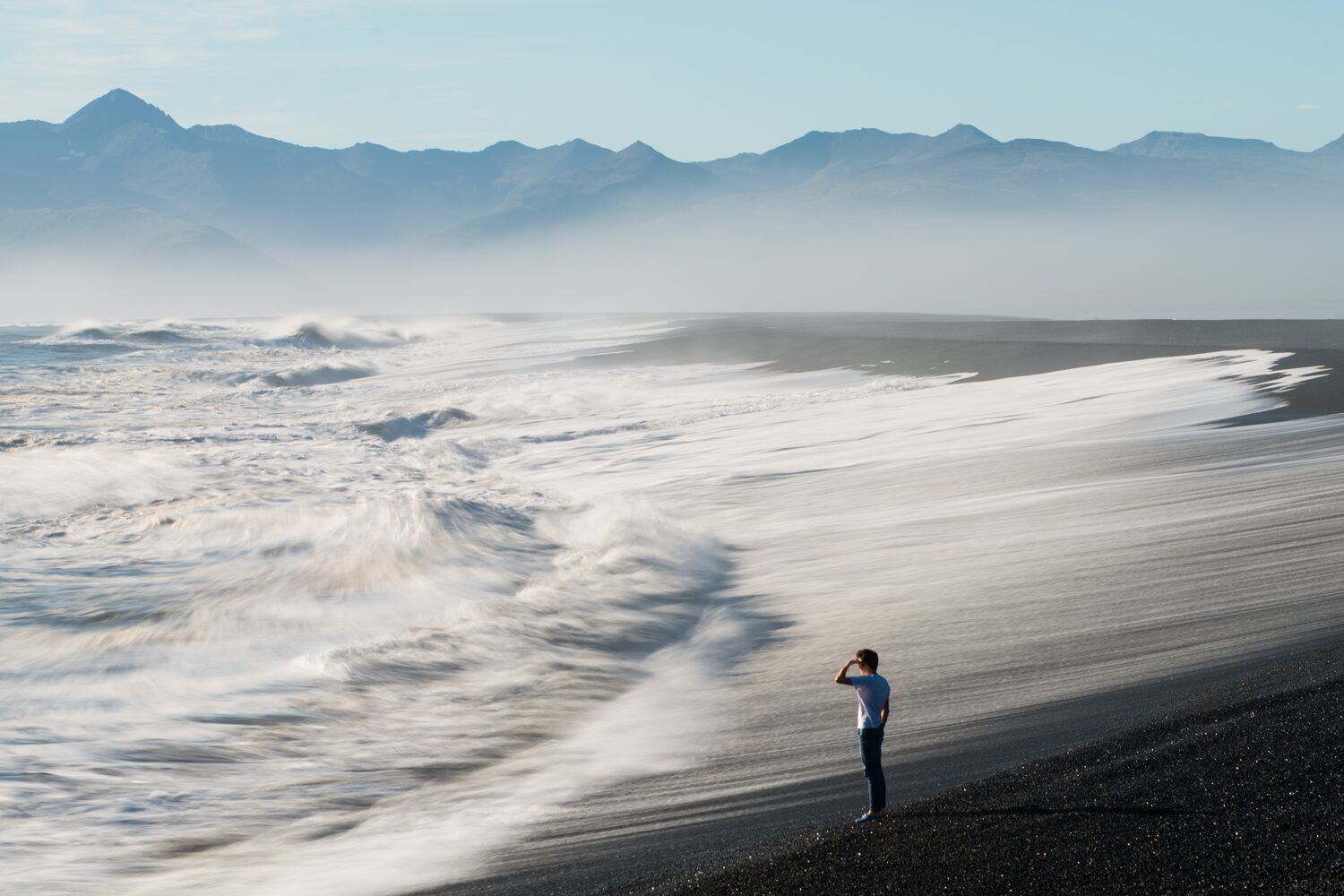 Man standing on black sand beach, Iceland, staring out at crashing ocean waves, with mountains in background.