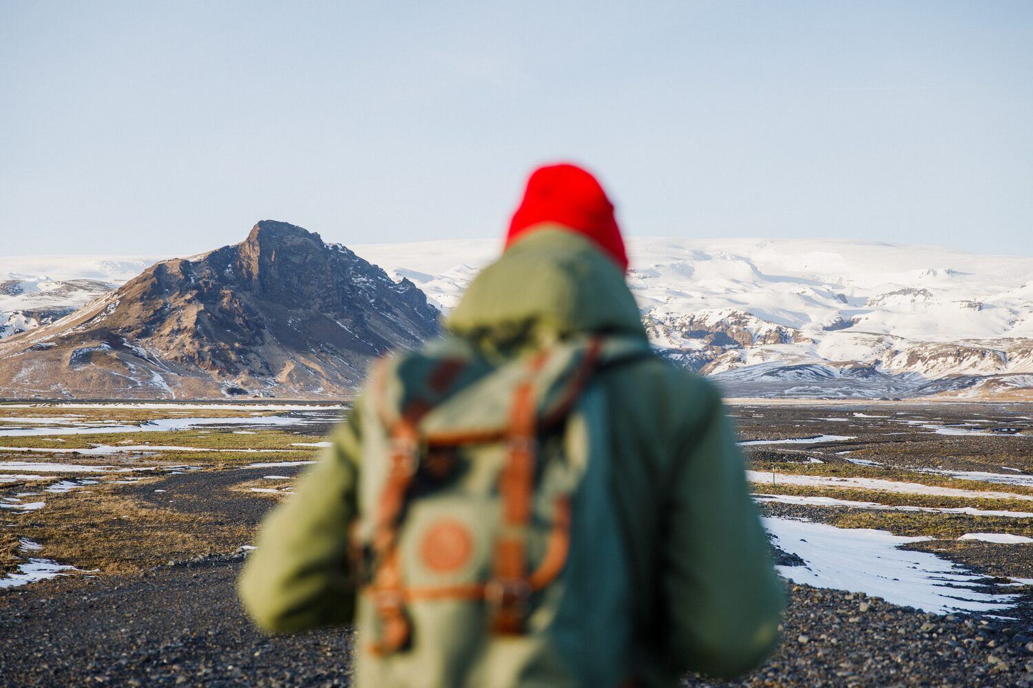 Male traveler in green coat and red beanie hat with green backpack, walking on cobbled path, with Icelandic snowy mountains in background.