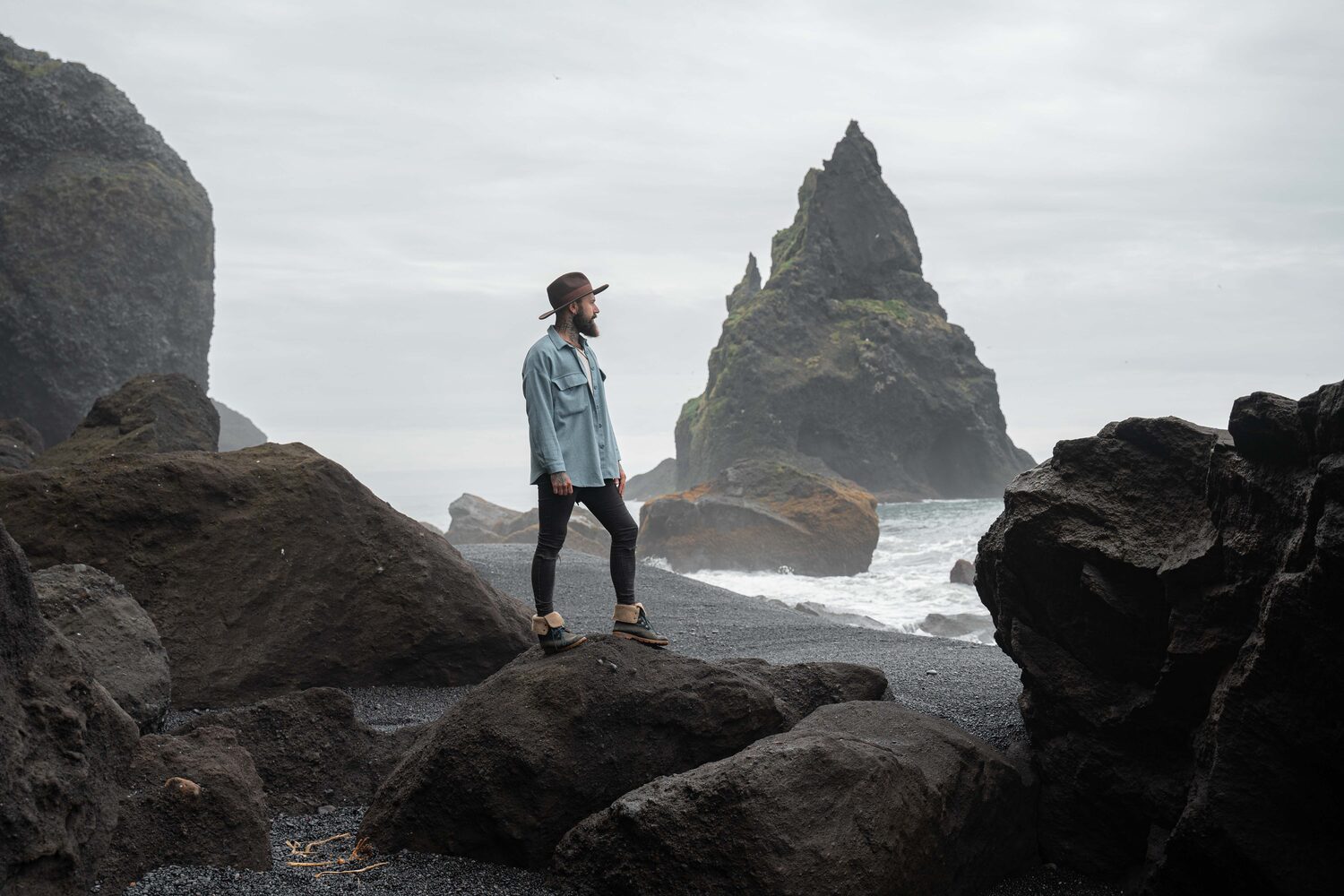 Powerful waves crashing over large rocks. View of man with bears in hat standing on rock and looking into the distance of the sea. 