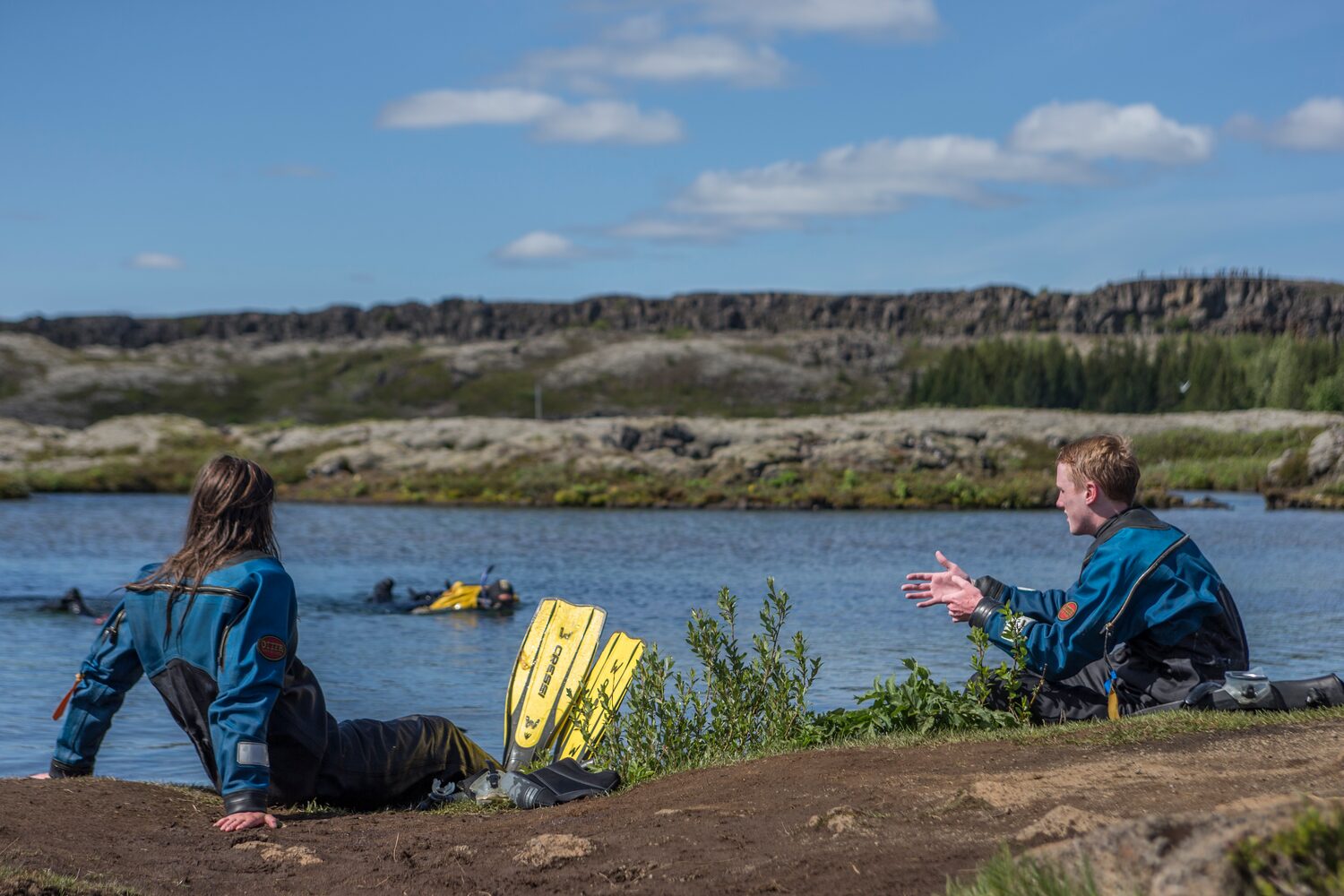 Silfra fissure snorkeling, two tourists dressed in snorkeling gear resting next to lake.