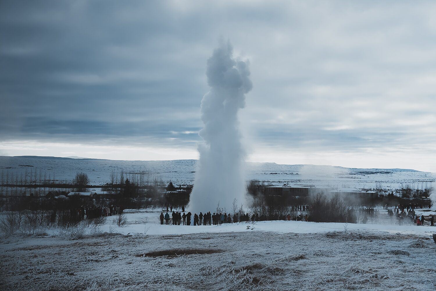 Large Geysir with smoke, in winter landscape with crowd of tourists watching with frozen ground in foreground 