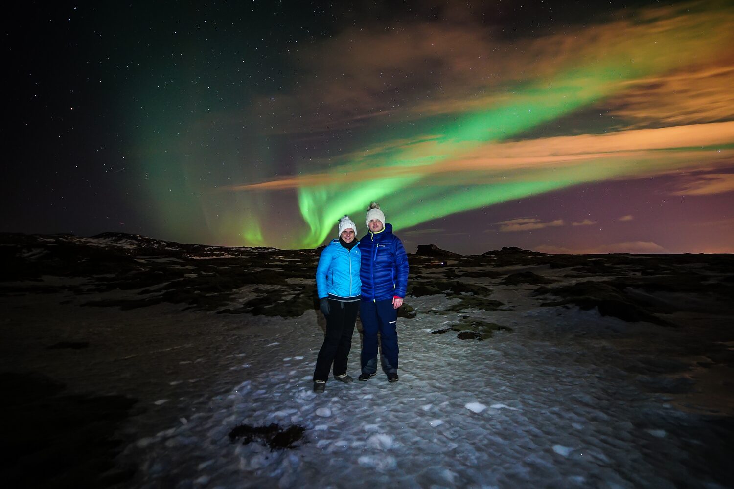 A couple in winter jackets and white beanies hugging while standing on snow under green and purple northern lights with clouds