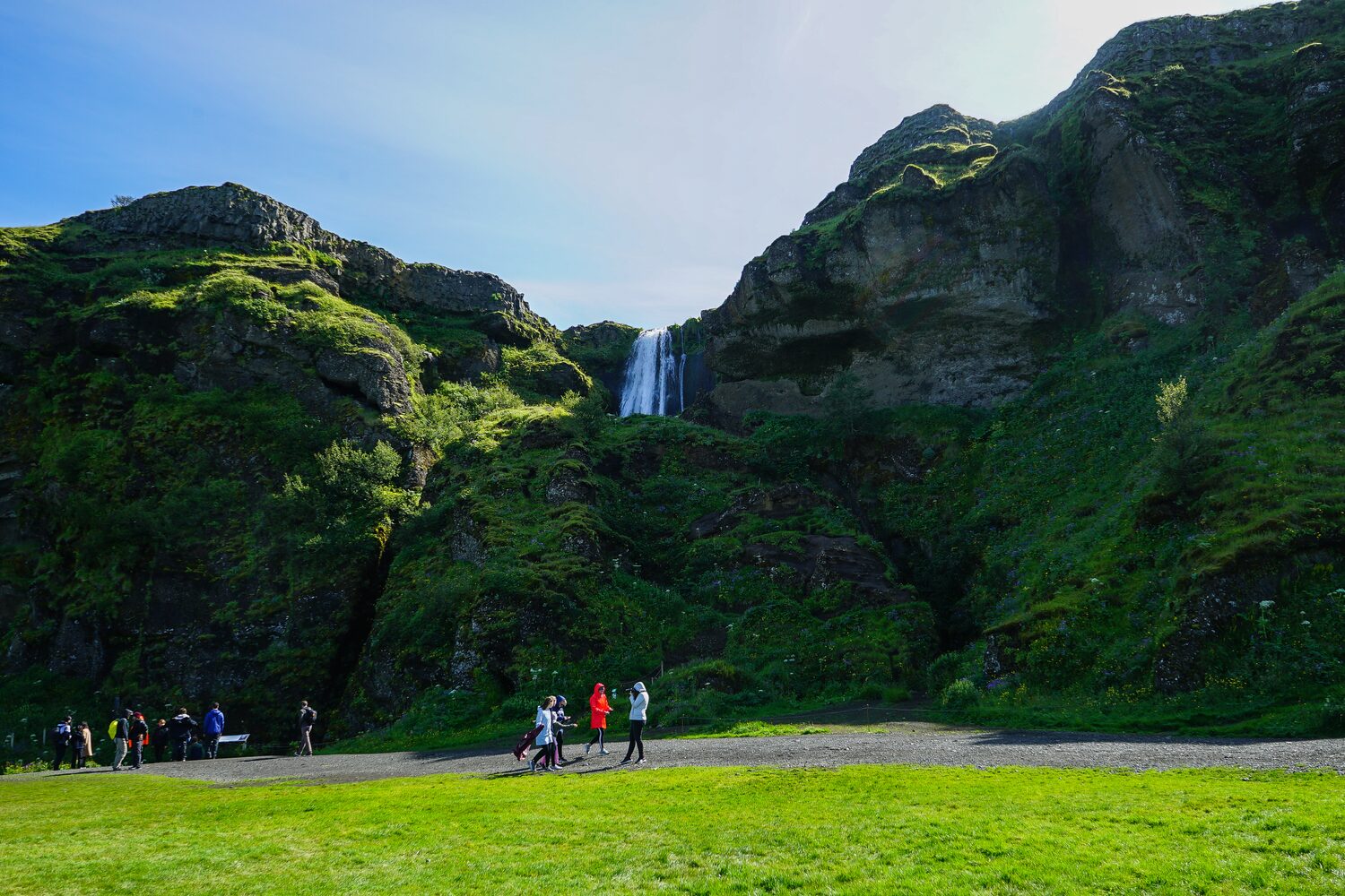 People walking on path below large mossy cliff with small waterfall in center