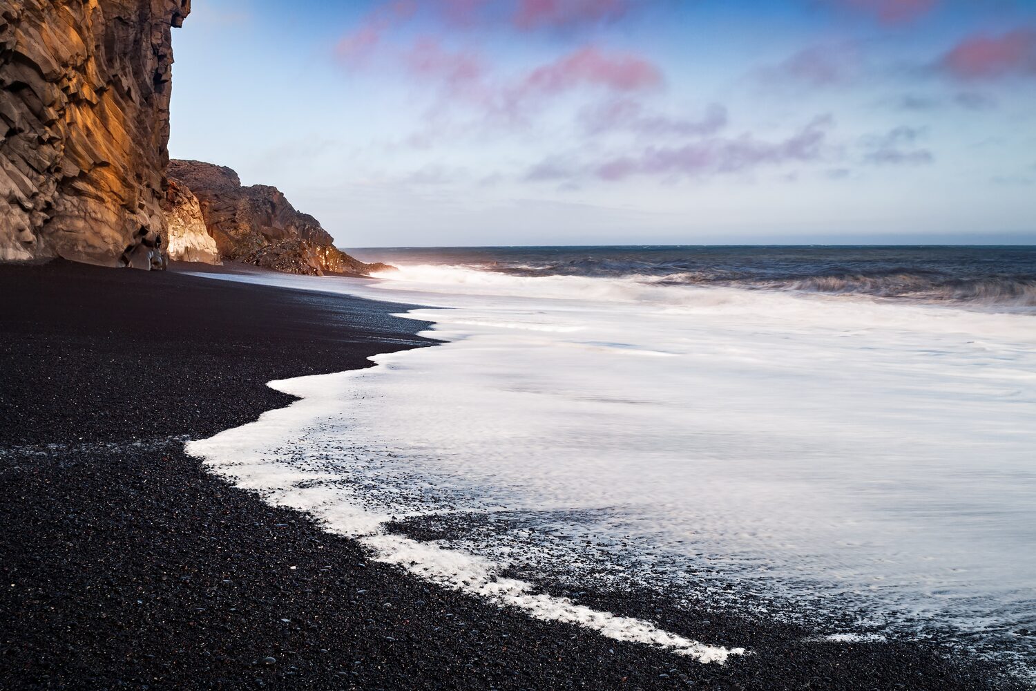 Blank pebbled beach with frothy white waves crashing shore and cliffs in distance with pink clouds 