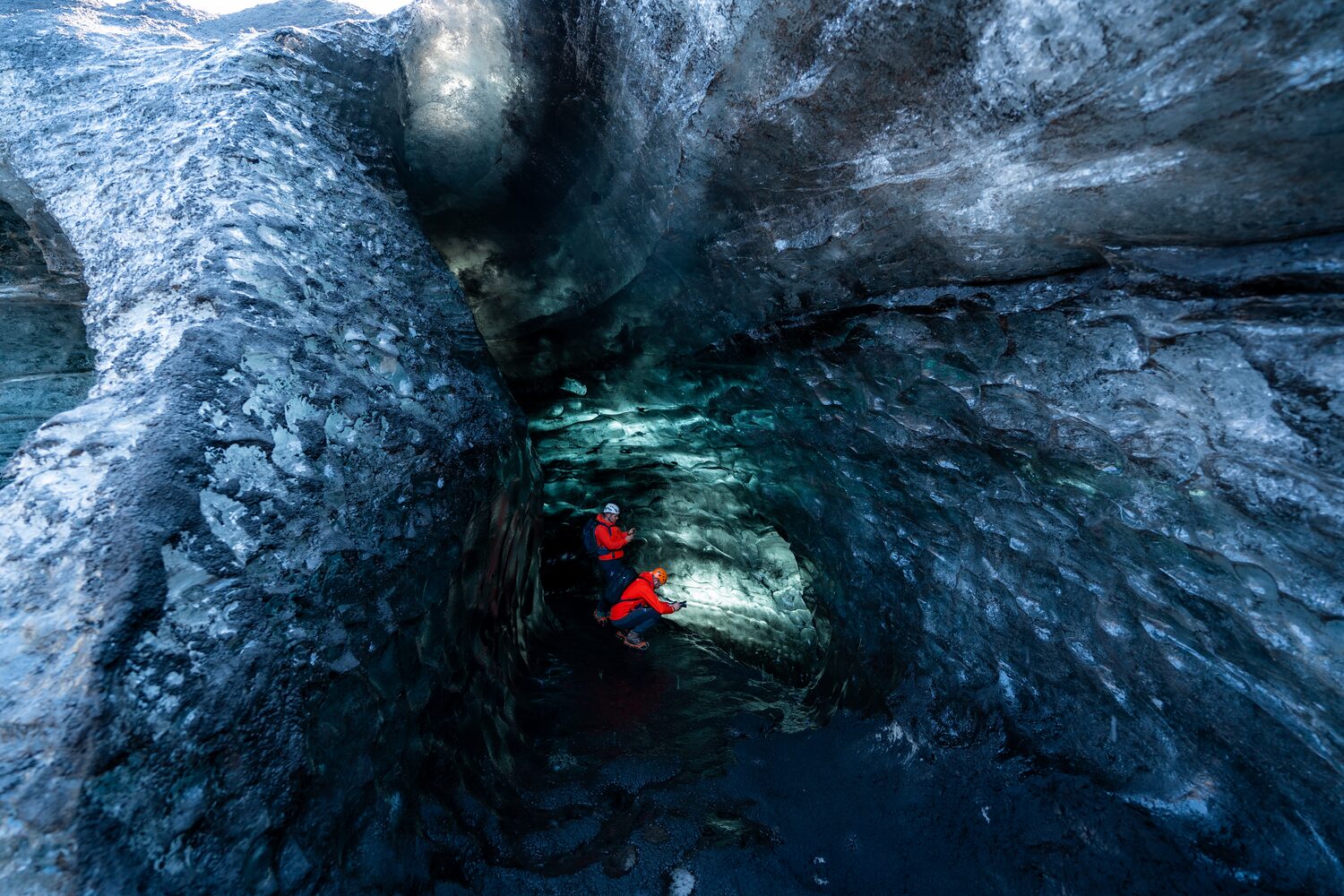 Two tourists in red coats looking at the beautiful patterns from inside an ice cave bright blue textured walls with reflecting light