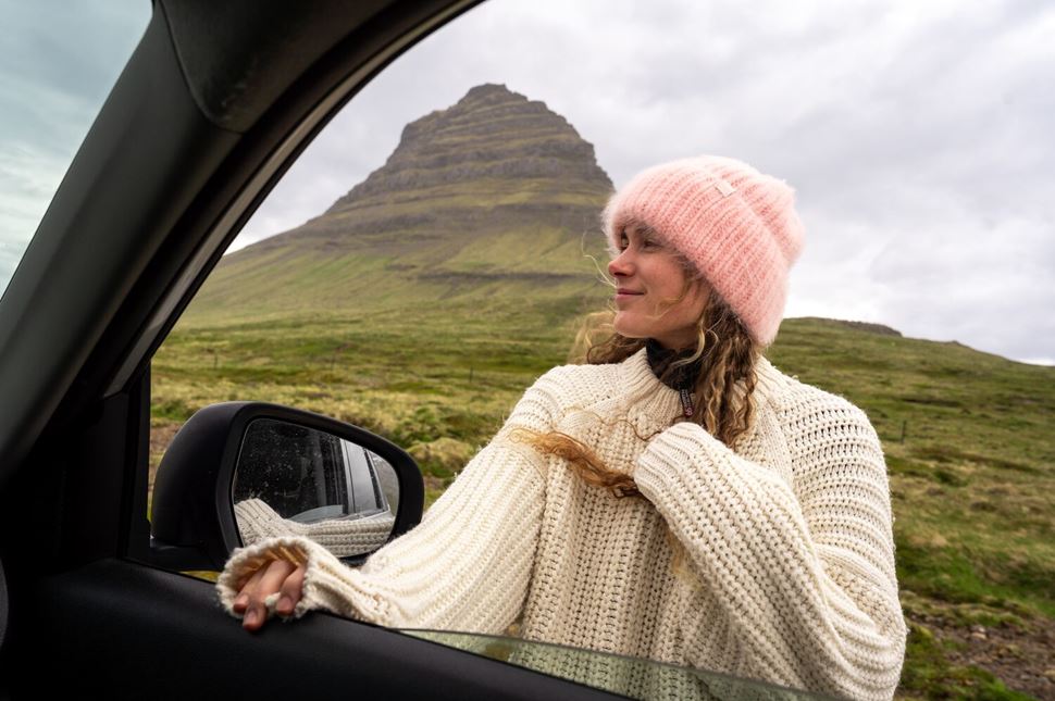 Woman Car Kirkufjell Woman dressed in autumn pink hat and jumper standing outside of car by Kirkjufell green hill