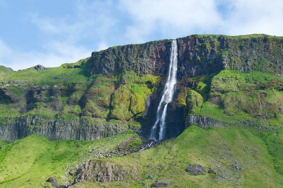 Peninsula Bjarnafoss Waterfall coming from tall cliff top covered in green grass bright blue skies with clouds