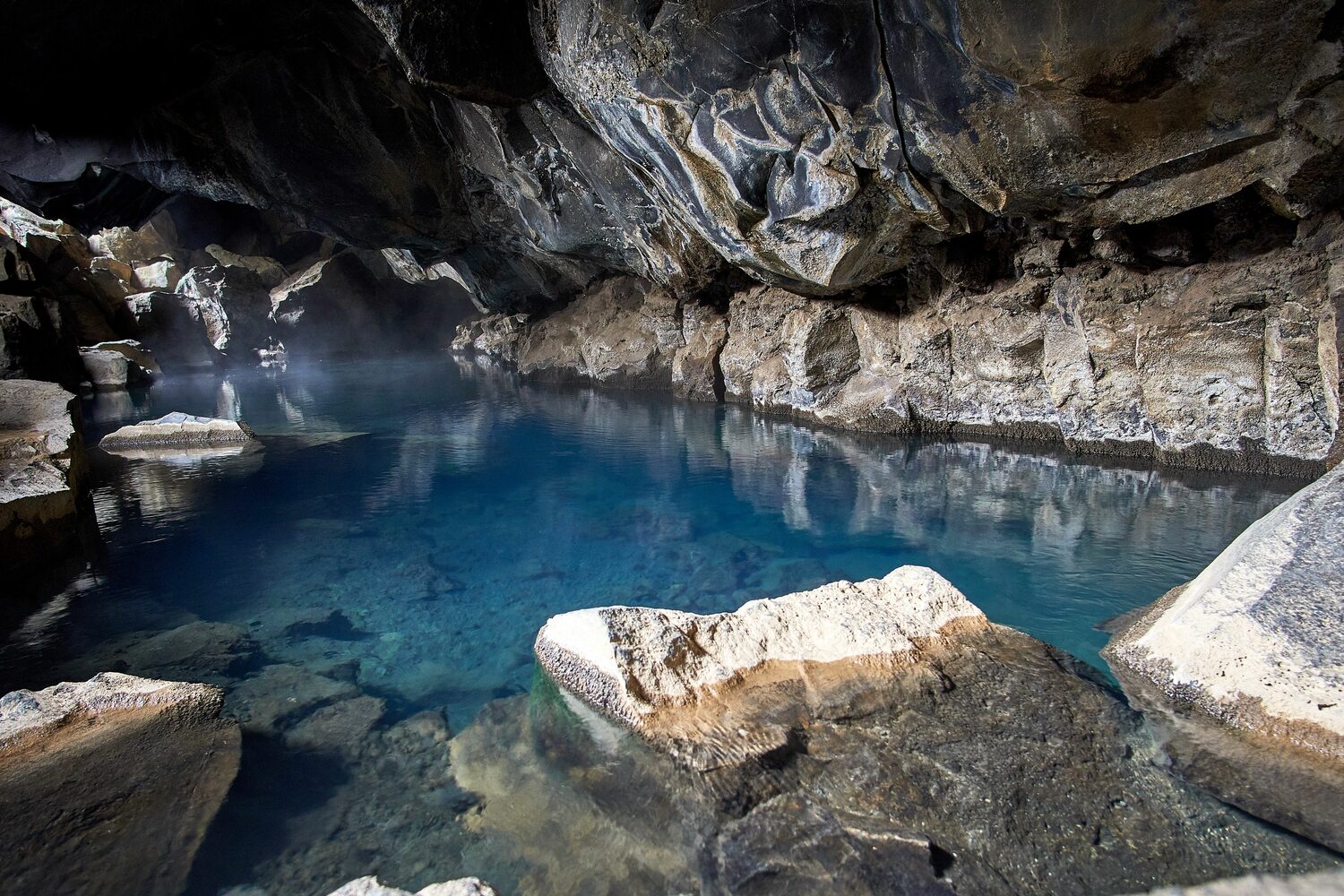 Inside of lava cave with large rocks and clear dark blue natural lake inside with natural light shining through