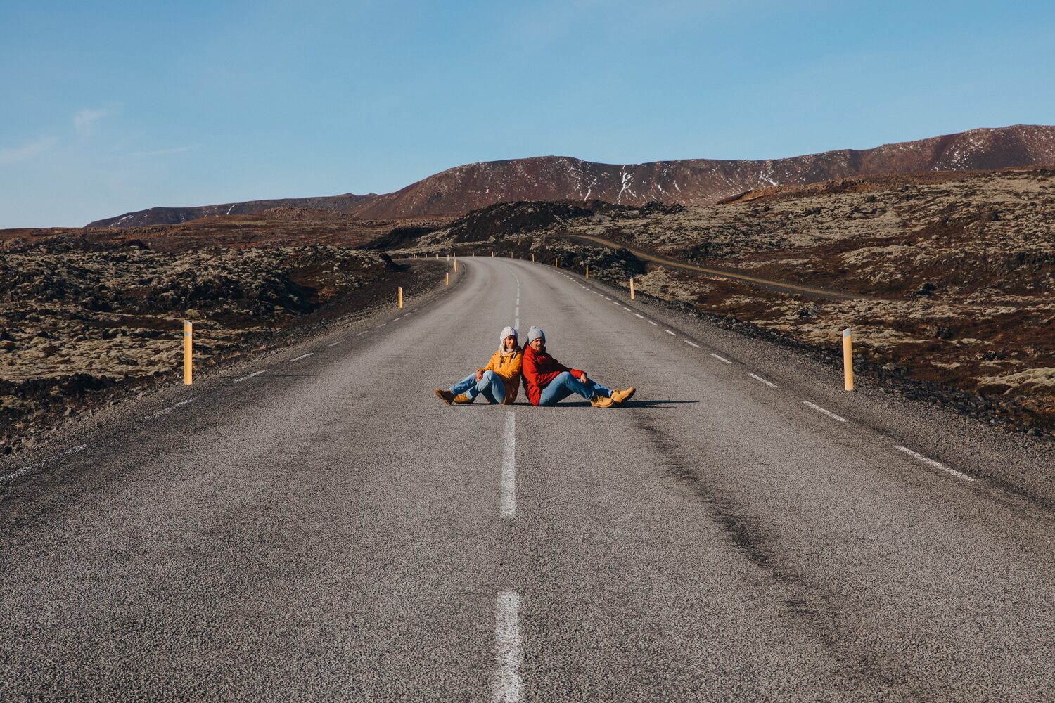 Couple sitting in middle of road back to back posing for photo in front of hilly landscape