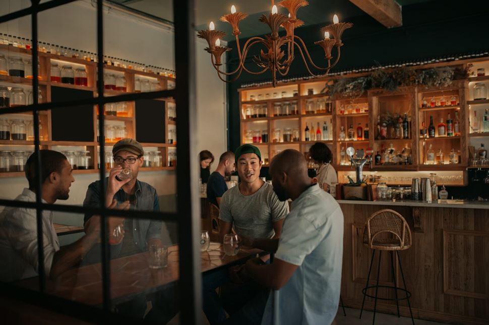 Male Friend Group Bar Group of four male friends sitting around table talking and drinking bar behind them with drinks on shelves