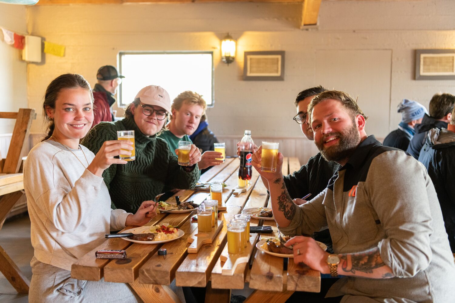 Group of five friends sitting at wooden table with plates of food and beers showing cheers for photo