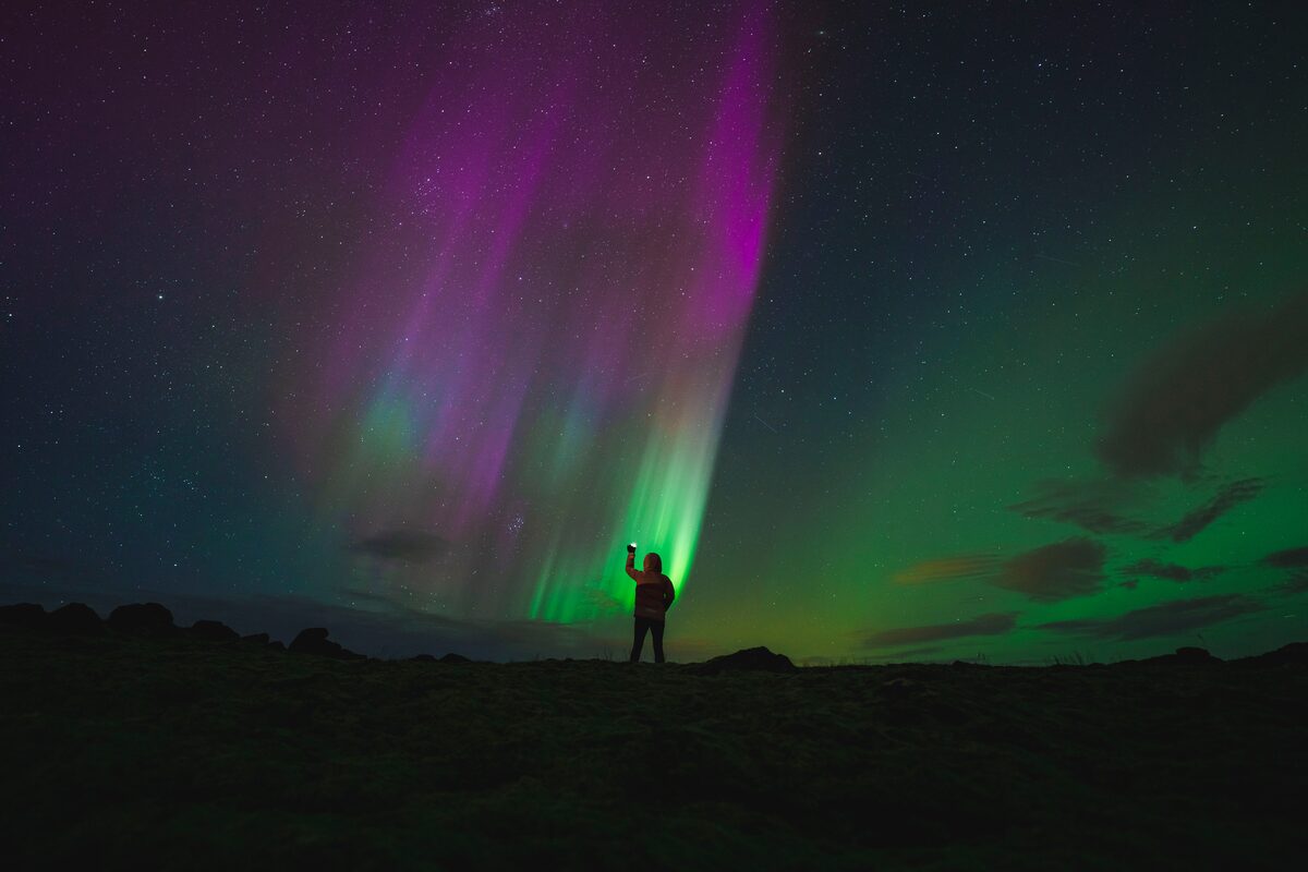 Tourist standing holding light up to sky staring at pink and green aurora colors