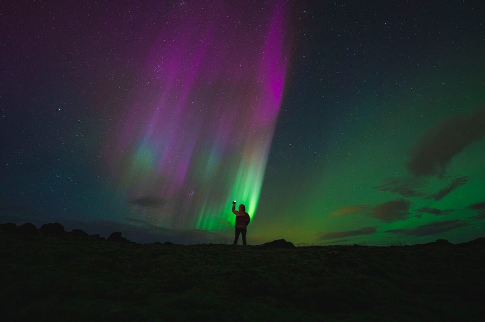 Tourist standing holding light up to sky staring at pink and green aurora colors