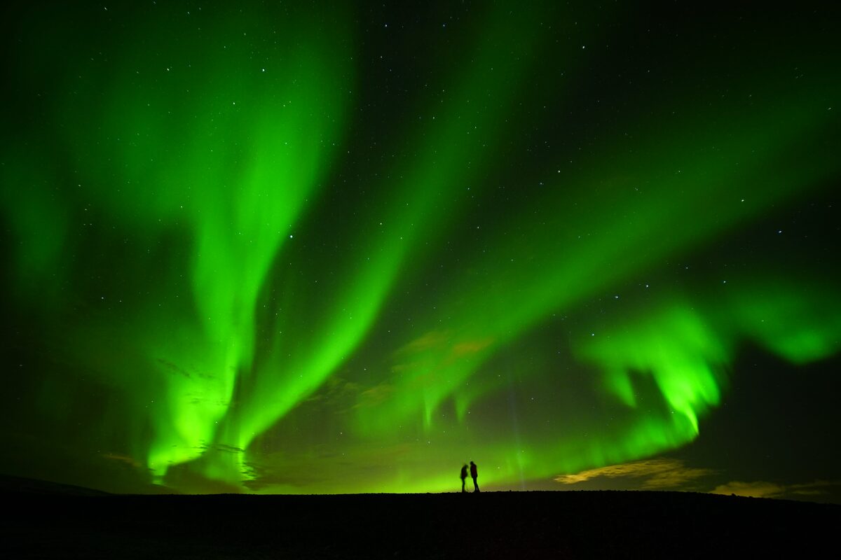 Two people standing underneath green northern lights in night sky.