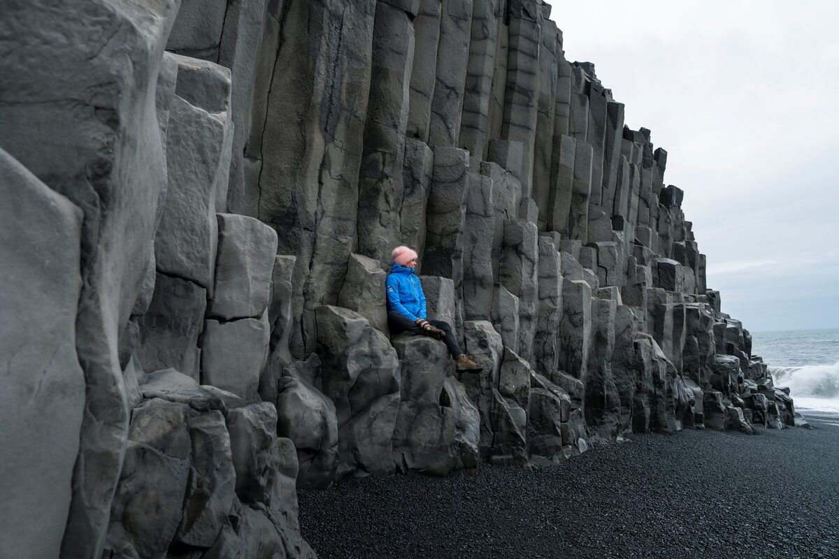Tourist in blue coat sitting up on Reynisfjara sea stacks looking at black sand beach 