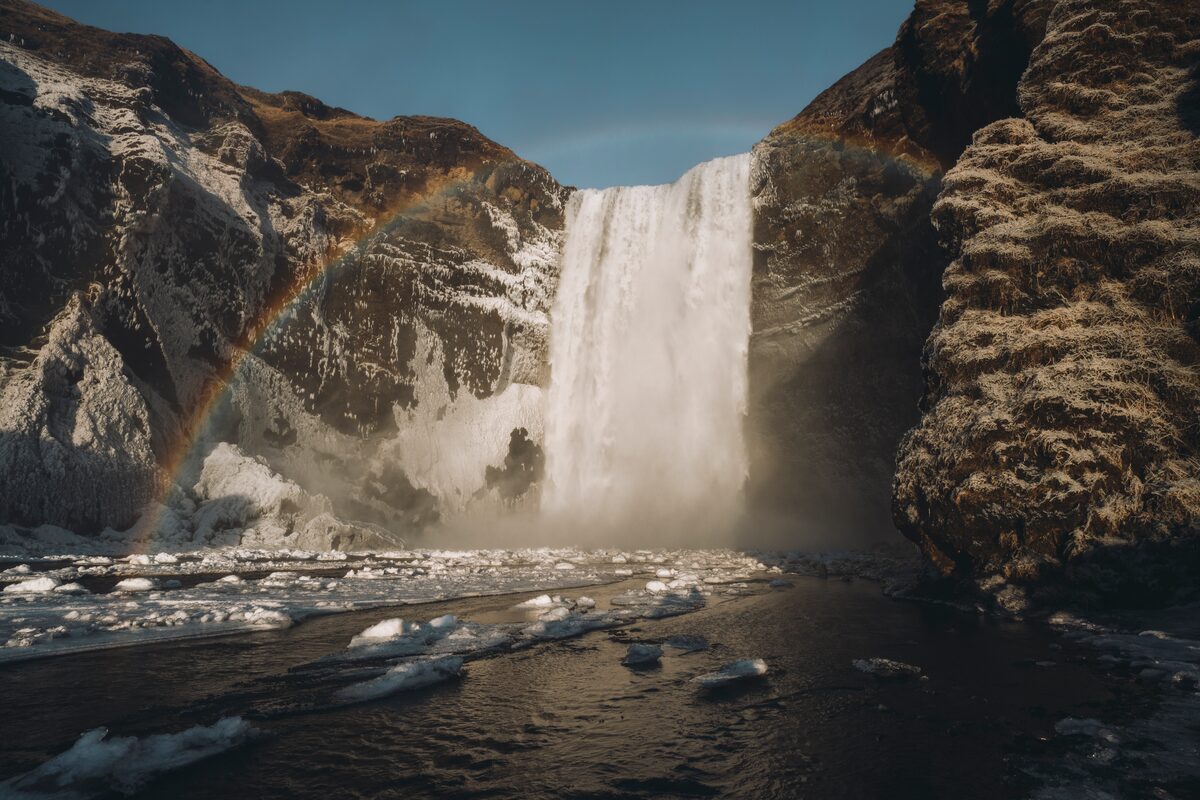 Snowy cliffs with icy waterfall flowing into snow covered river with rainbow 