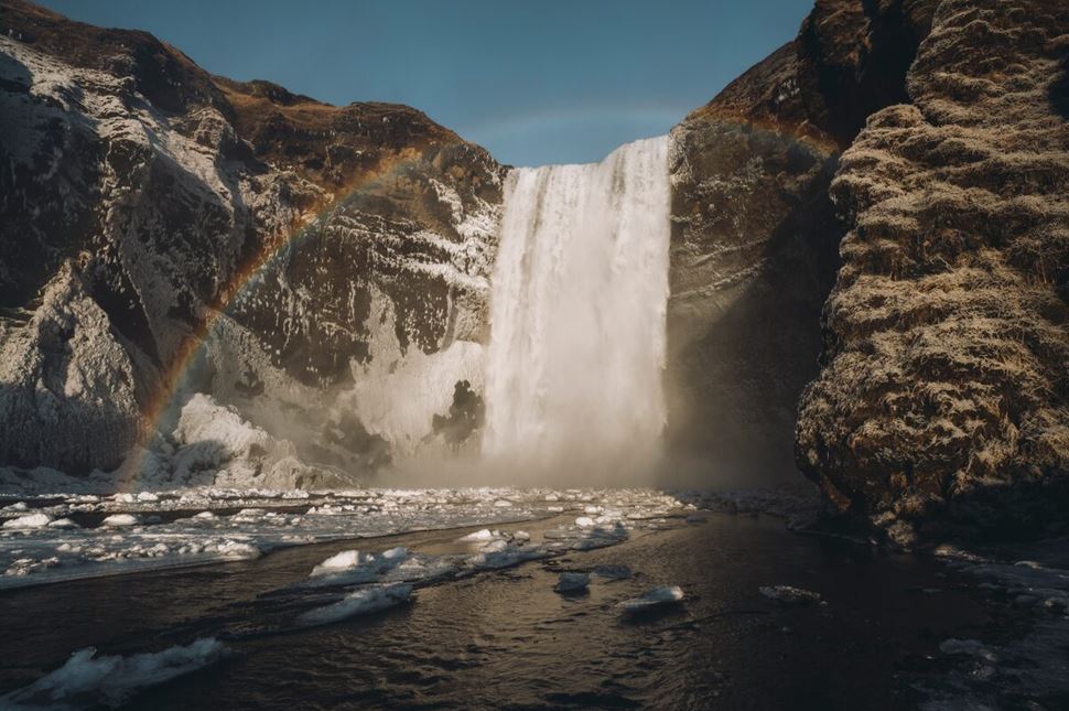 Snowy cliffs with icy waterfall flowing into snow covered river with rainbow