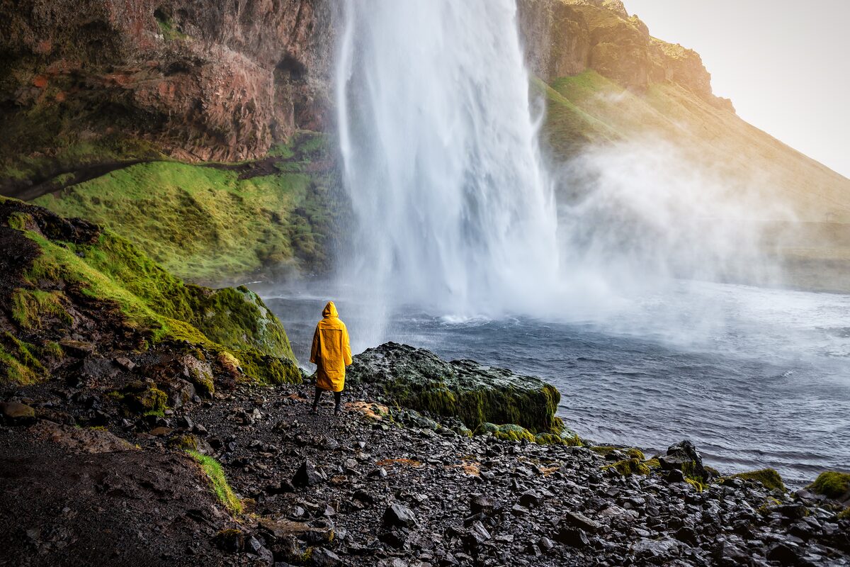 Female tourist standing underneath Seljalandsfoss waterfall in long yellow raincoat