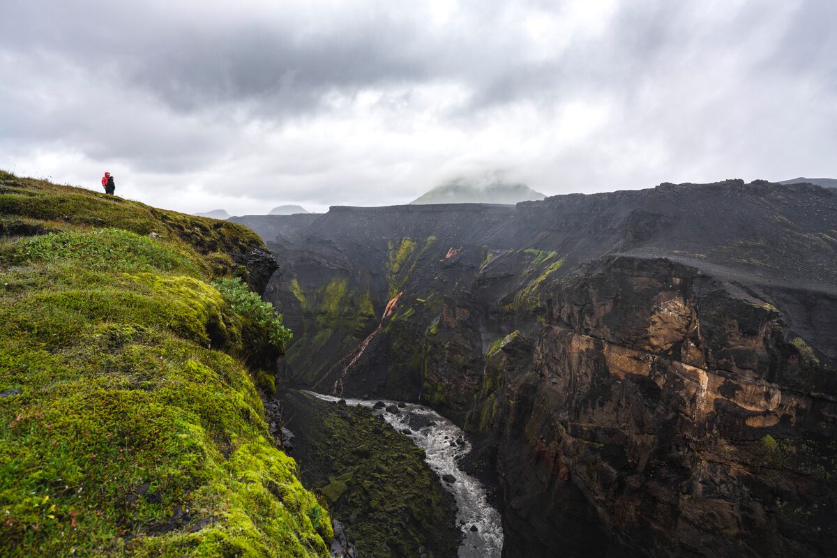 Two tourists standing at the edge of Markarfljotsgljufur Canyon covered in green icelandic moss