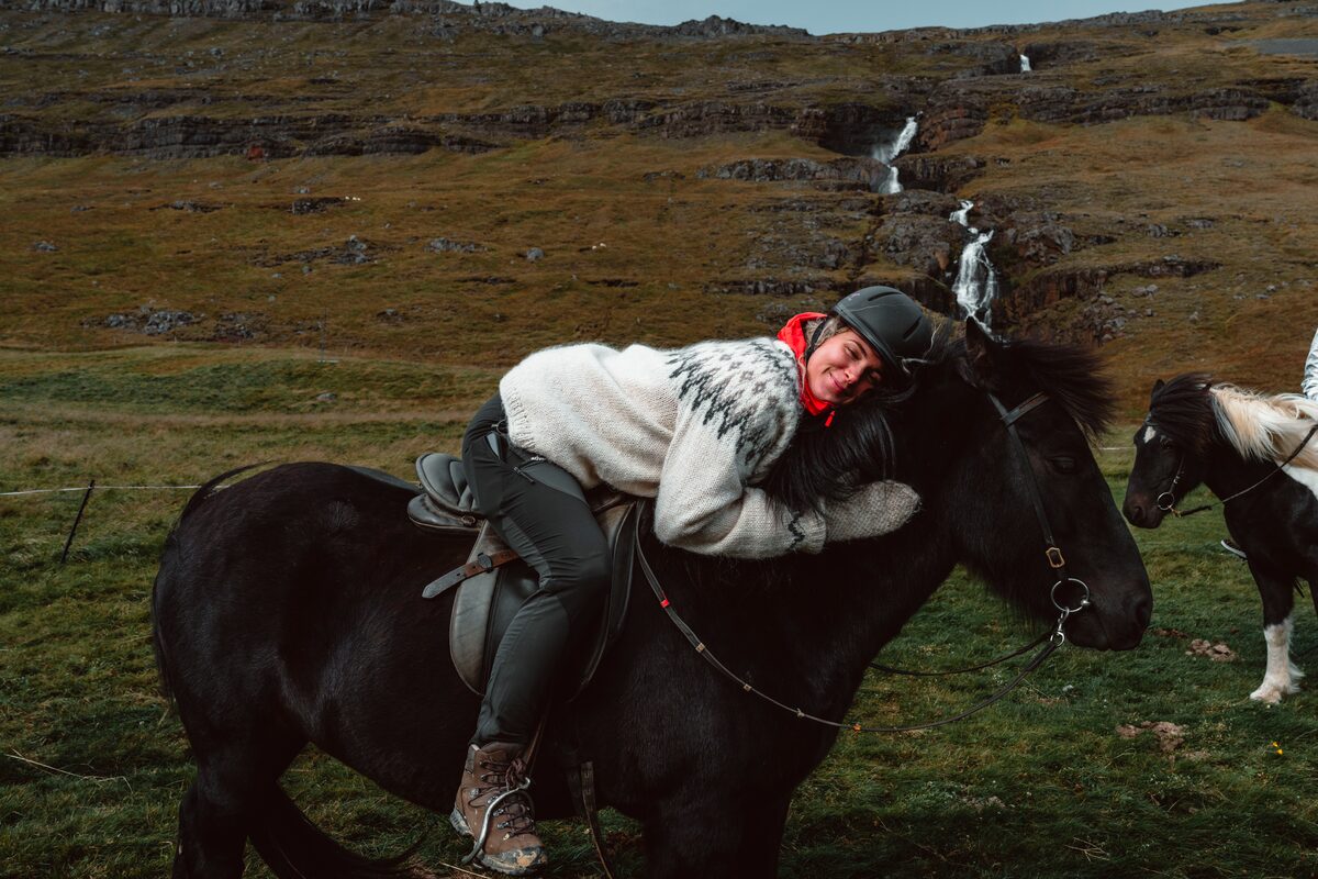 Lady wering icelandic jumper sitting on black horse hugging round horses neck 