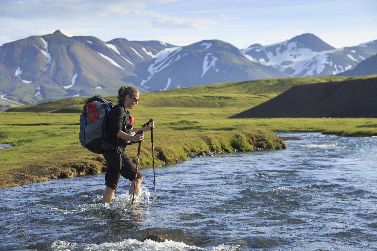 Lady with large hiking backpack and hiking sticks crossing stream Landmannalaugar